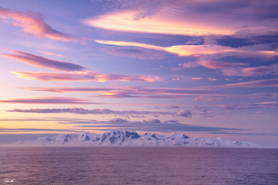 A photograph of Antarctica with a colorful sunset and snow covering the distant mountains across the Drake Passage