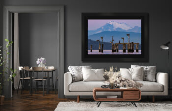 Large dark framed fine art print of a snow-capped volcano by the ocean at sunset displayed in the living room of a modern home